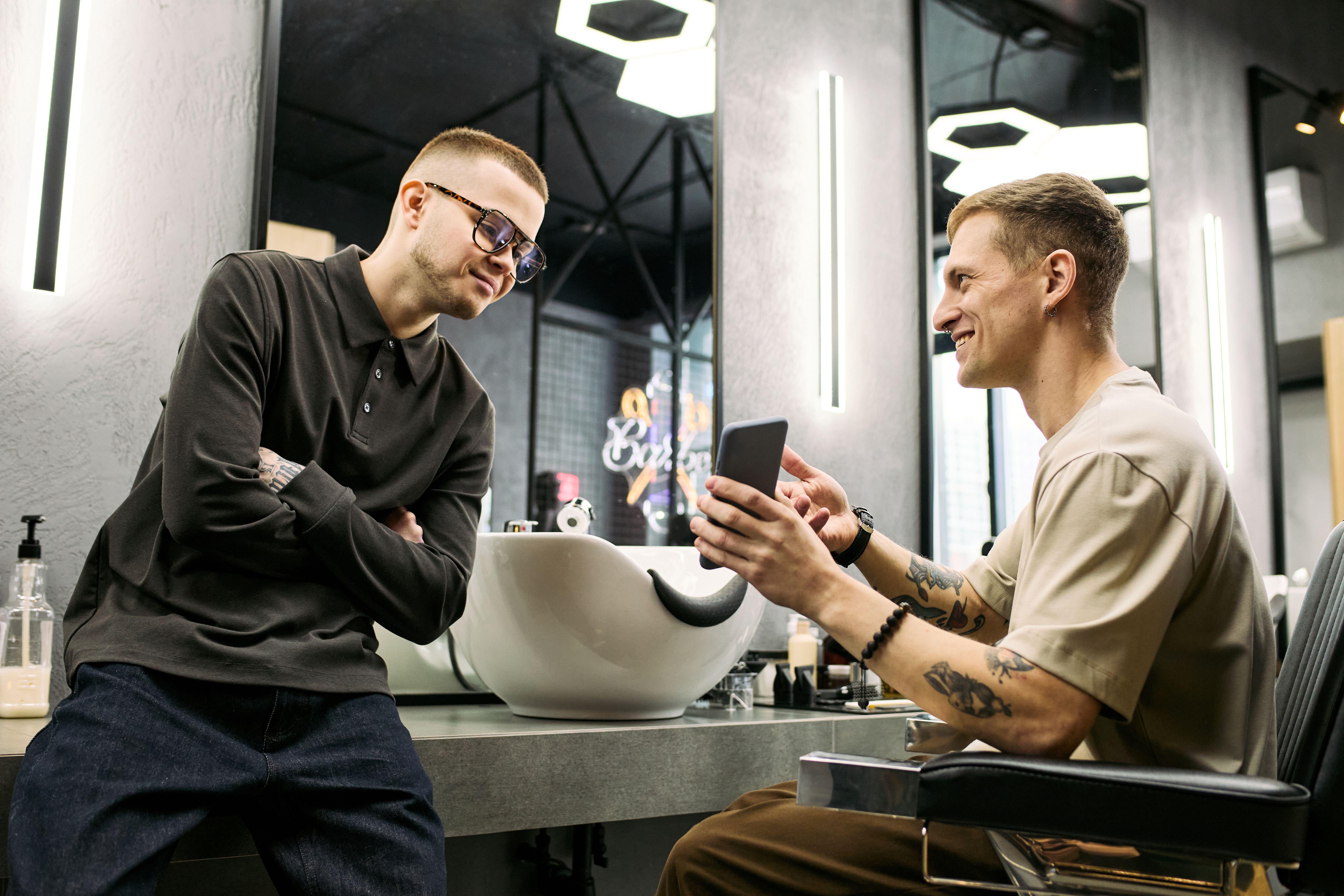 Zwei junge Männer im Friseursalon, einer zeigt dem anderen etwas auf dem Smartphone